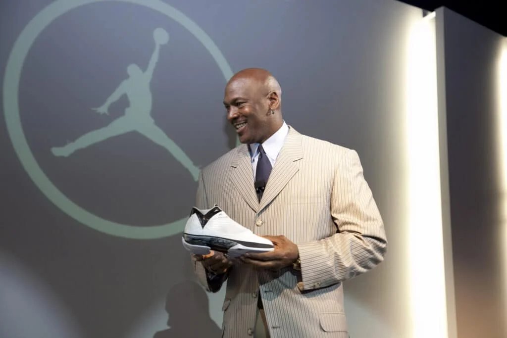A man in a striped suit is smiling and holding a white sneaker, standing in front of a large Jordan logo projected on the wall.
