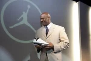 A man in a striped suit is smiling and holding a white sneaker, standing in front of a large Jordan logo projected on the wall.