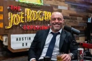 A man in a suit and tie sits smiling at a podcast desk, holding a glass of drink with a microphone in front of him and a neon sign reading "The Joe Rogan Experience" in the background.