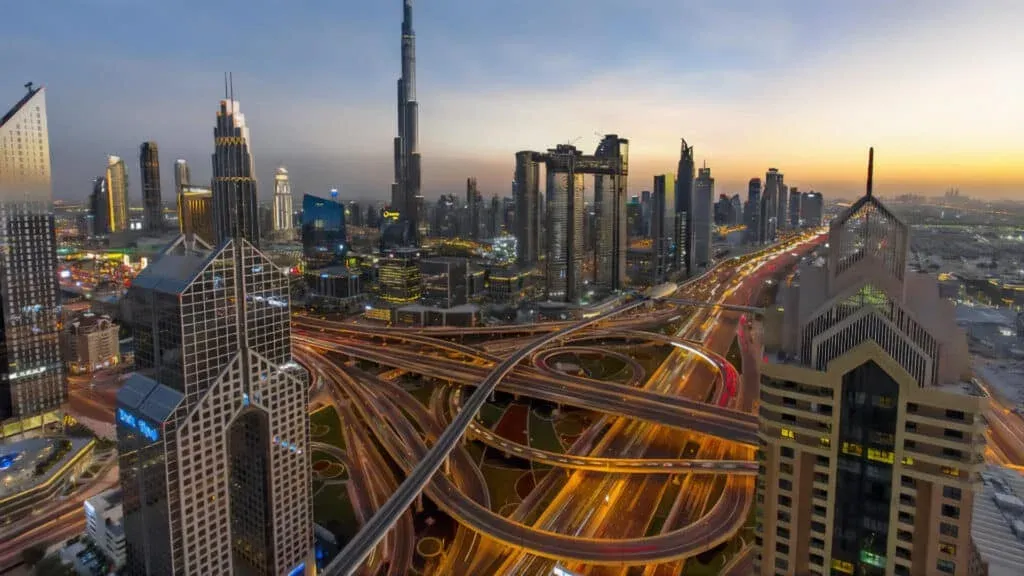 Aerial view of Dubai's skyline at dusk, featuring illuminated skyscrapers and intricate highway interchanges.