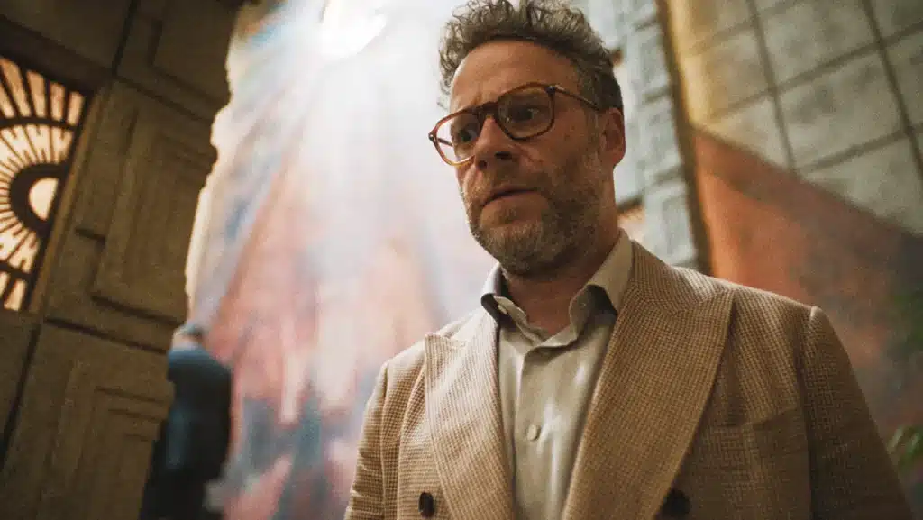 A man with glasses and a beard wearing a beige checkered blazer is standing indoors in front of a textured wall with light streaming in from above.