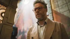 A man with glasses and a beard wearing a beige checkered blazer is standing indoors in front of a textured wall with light streaming in from above.