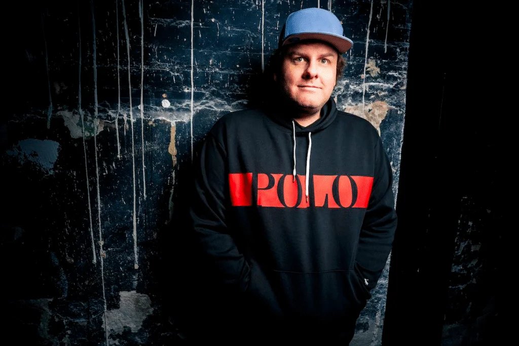 A person wearing a blue cap and a black hoodie with "POLO" written in red stands against a textured, dark blue wall with white streaks.