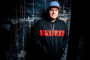 A person wearing a blue cap and a black hoodie with "POLO" written in red stands against a textured, dark blue wall with white streaks.