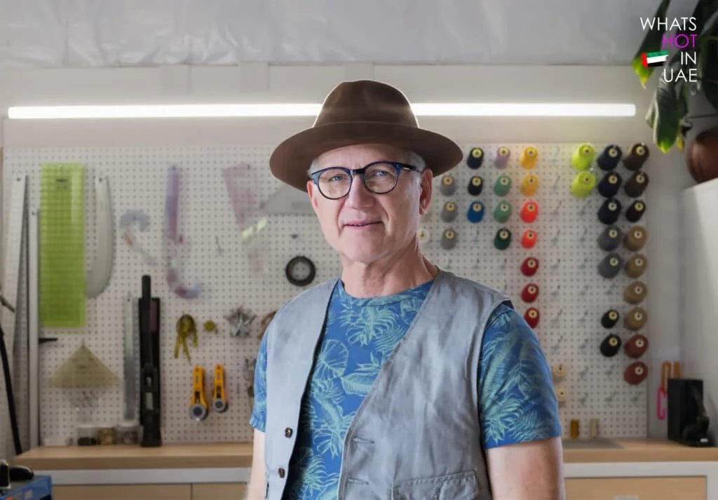 A person wearing a brown hat, glasses, and a blue tropical print shirt with a gray vest stands in a workshop with a pegboard displaying various colorful spools and tools.