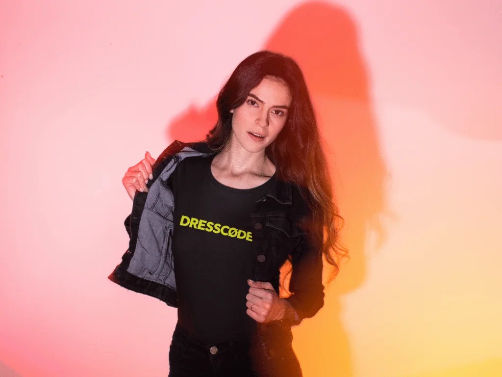 A person with long hair poses against a pink and orange backdrop, wearing a black denim jacket over a black shirt with the word "DRESSCODE" in yellow letters.