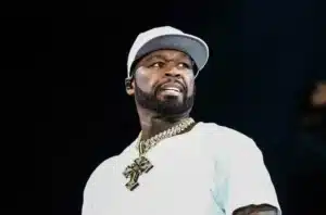 A man wearing a white cap and shirt, with a prominent beard and a large gold chain visible around his neck, looking slightly to the side with an expressive facial expression against a dark background.