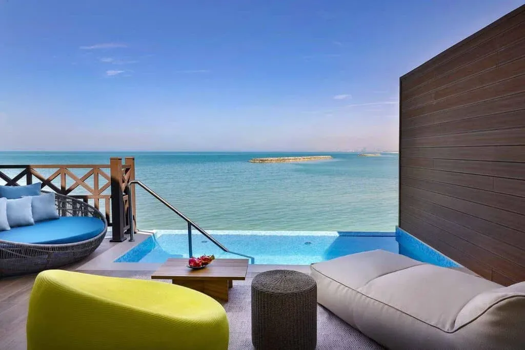 A luxurious outdoor seating area overlooking a tranquil sea, featuring a blue circular lounge chair, a yellow accent chair, and a small wooden table with decorative fruits, alongside a plunge pool.