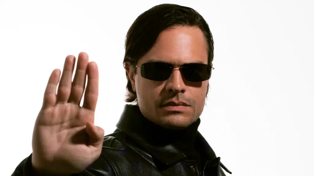 A man wearing sunglasses and a black leather jacket stands against a white background, holding his hand up in a stop gesture.