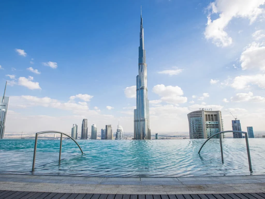 A panoramic view of the Burj Khalifa towering over a skyline dotted with modern skyscrapers, with an infinity pool in the foreground reflecting the clear blue sky and fluffy clouds.