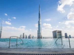 A panoramic view of the Burj Khalifa towering over a skyline dotted with modern skyscrapers, with an infinity pool in the foreground reflecting the clear blue sky and fluffy clouds.