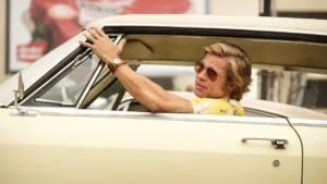 A man with blonde hair wearing sunglasses and a yellow floral shirt leans out of the driver's side window of a vintage yellow car, gesturing one hand while resting the other on the window frame.