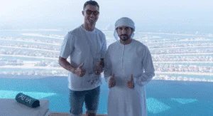 Two men posing with thumbs up next to an infinity pool, with a scenic view of the Palm Jumeirah in Dubai in the background. One man is wearing sunglasses and a casual T-shirt, while the other is dressed in traditional Emirati attire.