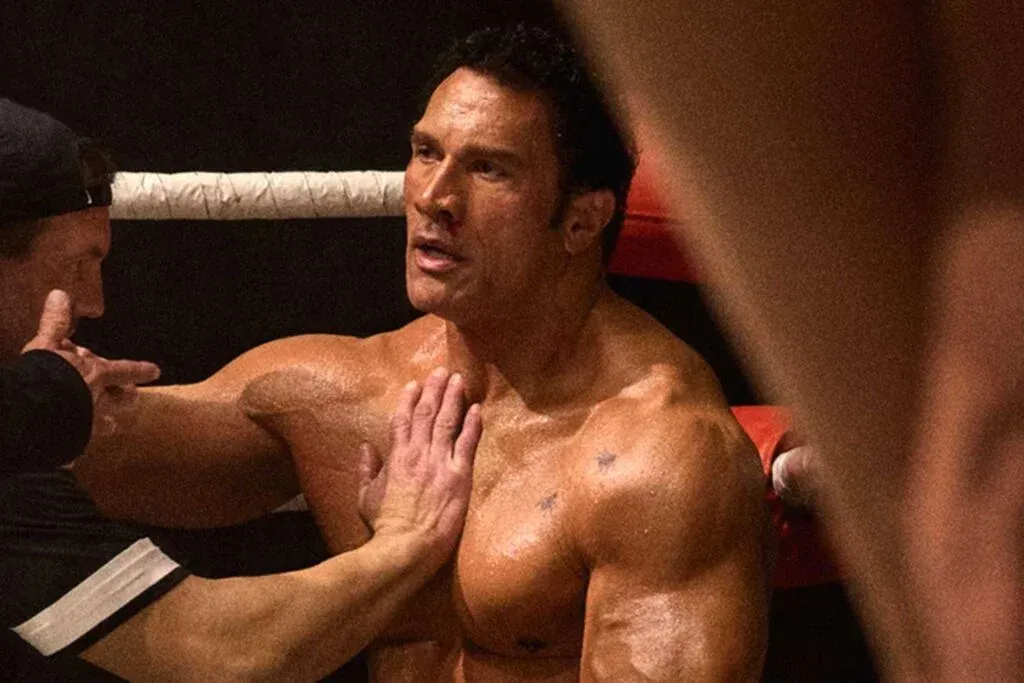 A muscular man with dark hair, shirtless and glistening with sweat, is receiving coaching from a trainer in a boxing ring, with red corner padding visible in the background.