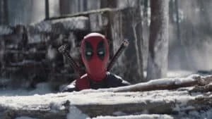 A masked figure in a red and black costume, wielding two swords on their back, peeks over a snow-covered log in a forest setting with trees in the background.