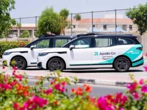 Two white autonomous vehicles branded with 'Apollo Go' are parked side by side in a sunny outdoor setting, with colorful flowers in the foreground and a fence in the background.
