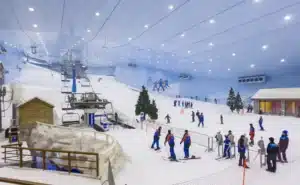 A spacious indoor ski facility featuring artificial snow, with a ski lift in the background and numerous skiers and snowboarders on the slopes. Surrounding areas include small trees and a rustic wooden structure.