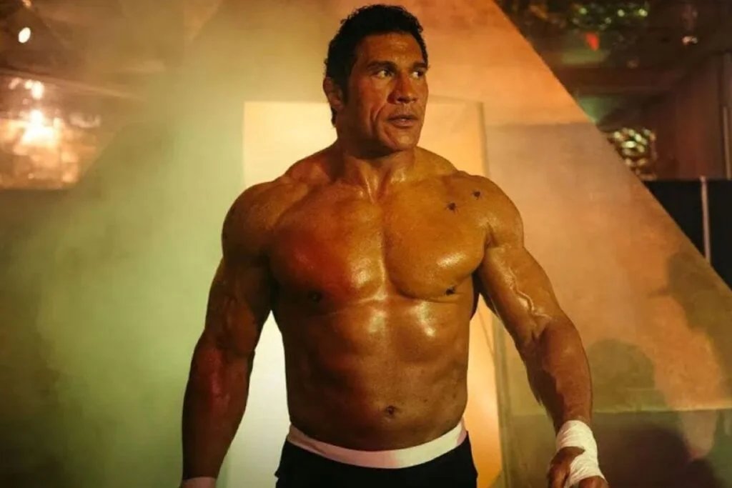 A muscular man with a tanned physique stands confidently in a misty, dimly lit environment, showcasing prominent muscles and wearing minimalist clothing.