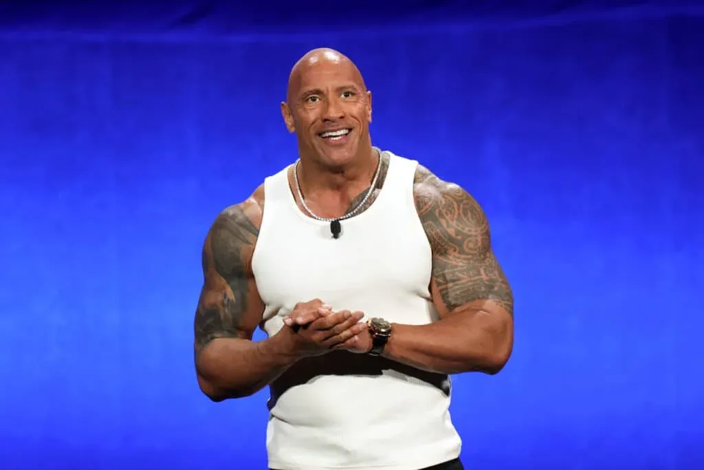 A muscular man wearing a white tank top stands in front of a blue background, smiling and gesturing with his hands.