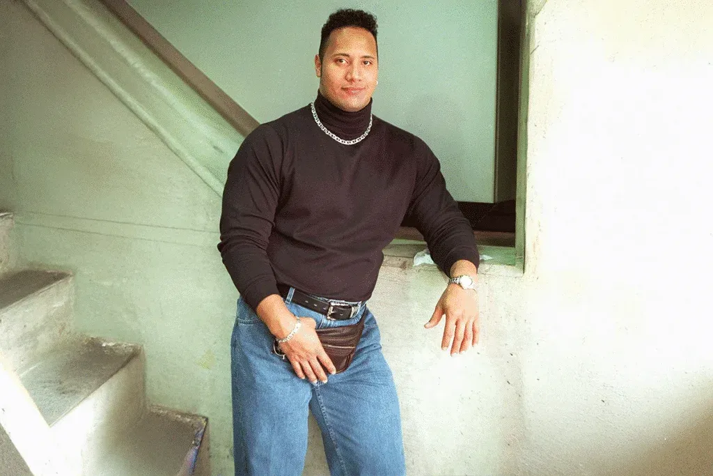 A muscular man stands in a casual pose against a wall, wearing a black turtleneck sweater, blue jeans, and a silver watch. He has short hair and is holding a small brown pouch. A staircase is visible in the background.