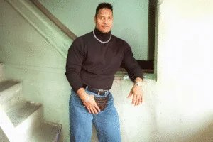 A muscular man stands in a casual pose against a wall, wearing a black turtleneck sweater, blue jeans, and a silver watch. He has short hair and is holding a small brown pouch. A staircase is visible in the background.