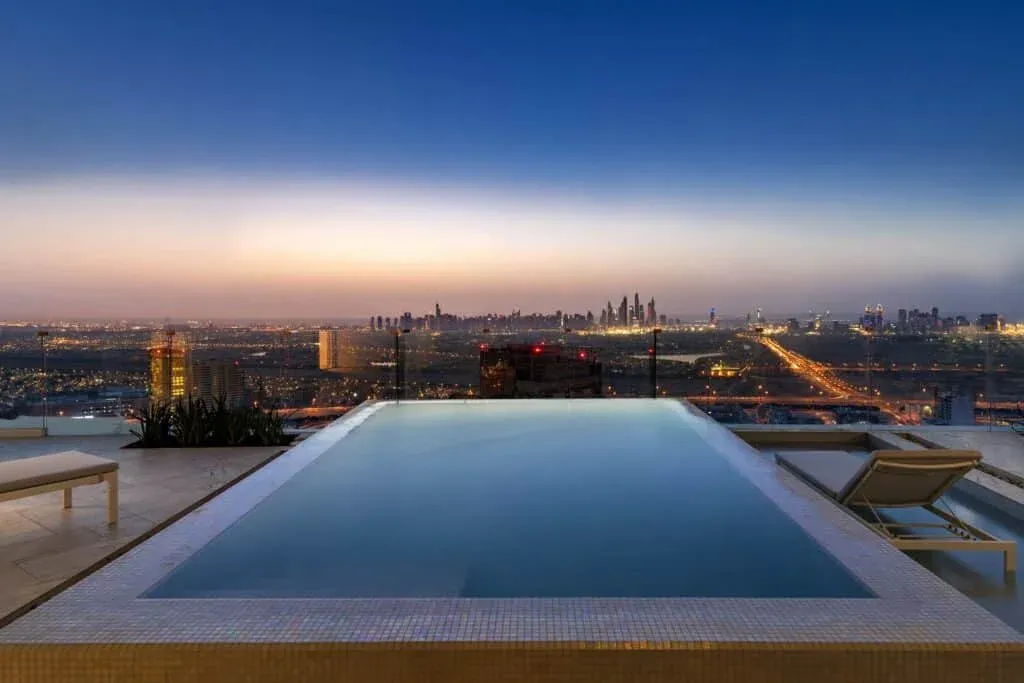 A rooftop infinity pool overlooking a city skyline at dusk, with lounge chairs placed nearby and city lights starting to illuminate the cityscape in the background.