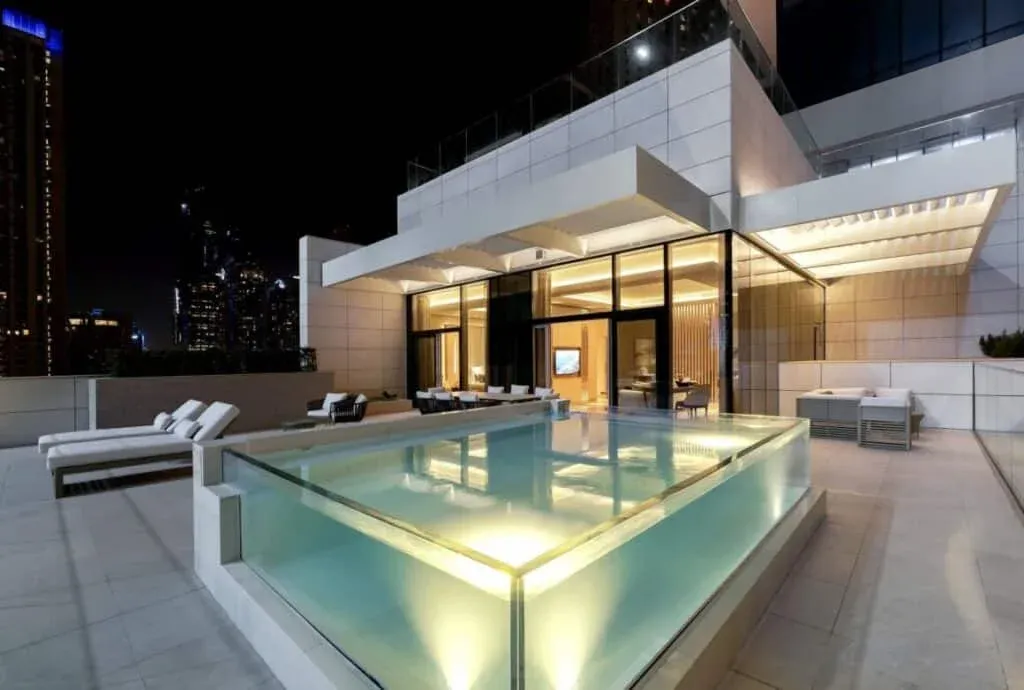 A modern rooftop terrace at night featuring a glass-sided pool, lounge chairs, and a stylish seating area, with city skyline lights in the background.