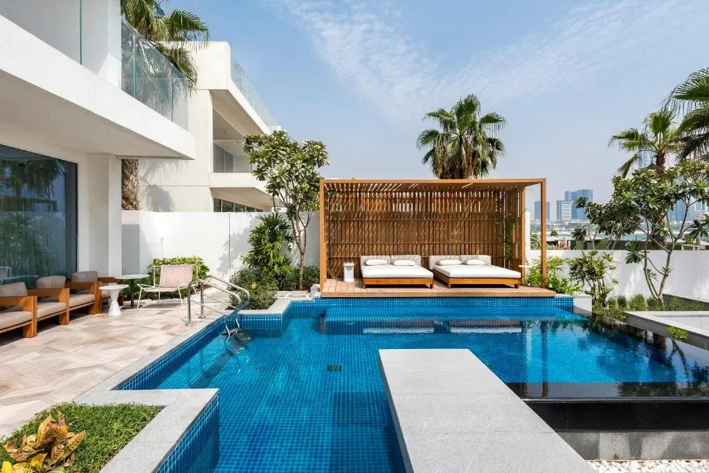 A modern outdoor pool area featuring a deep blue tiled pool with a wooden terrace and two white cushioned lounge beds under a bamboo shade structure, surrounded by lush greenery and palm trees, adjacent to a contemporary building with large windows.