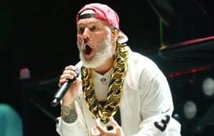 A performer with a white beard and a red cap shouts into a microphone while wearing a large gold chain necklace, dressed in a white jersey with NFL branding.