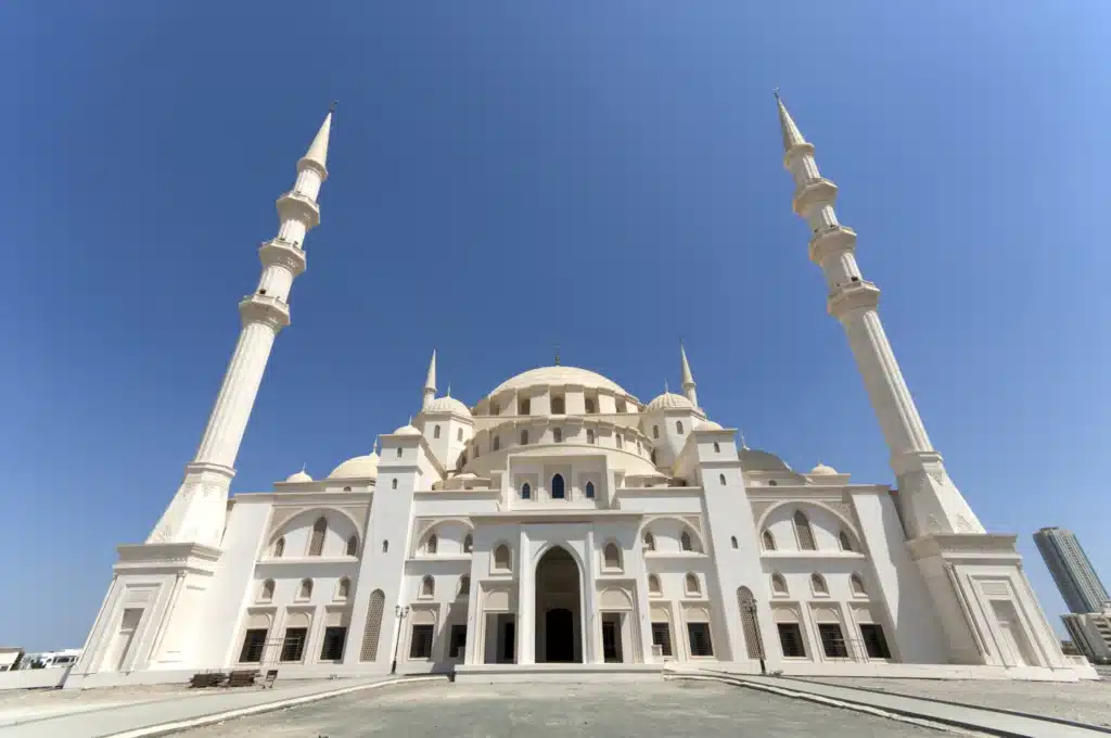 A grand white mosque featuring a large central dome and four tall minarets, set against a clear blue sky.