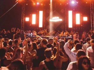 A lively crowd dancing at a music event, with vibrant stage lights and a DJ booth in the background, highlighting an energetic atmosphere filled with raised hands and diverse attendees.