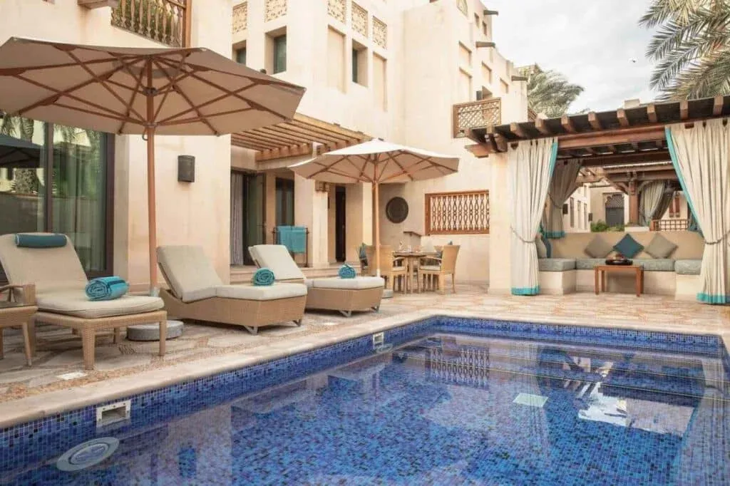 A luxurious outdoor space with a small blue tiled swimming pool surrounded by sun loungers with turquoise towels, large umbrellas for shade, and a cozy seating area draped with curtains. The setting features a backdrop of earthy-colored architecture and lush green palm trees.