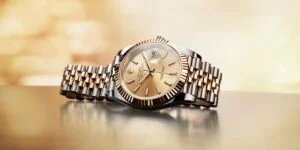 A luxury wristwatch with a champagne-colored dial, featuring a fluted bezel, a date window, and a two-tone metal bracelet combining gold and stainless steel, displayed on a reflective surface with a soft, blurred background.