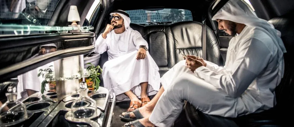 Two men in traditional white attire sitting inside a luxury limousine. One man is looking contemplatively out the window while the other is focused on his smartphone. The interior showcases plush leather seats and decorative elements.