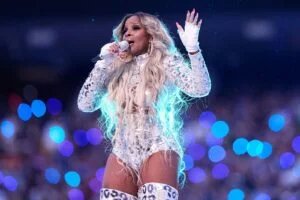 A performer on stage holds a microphone, wearing a glittery silver outfit with long blonde hair and dramatic makeup, illuminated by colorful lights in the background.