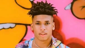 A young man with styled short hair and a colorful jacket stands in front of a vibrant pink and orange mural, looking directly at the camera with a serious expression.