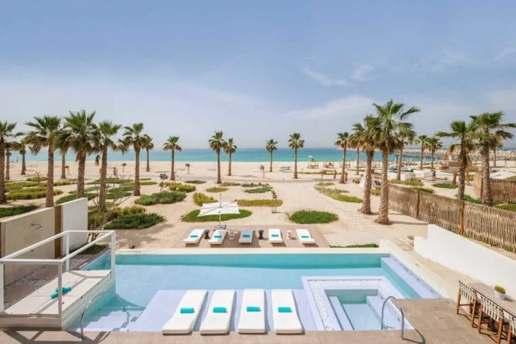 A luxurious outdoor pool overlooking a sandy beach with palm trees and the ocean in the background, featuring lounge chairs and shaded areas.