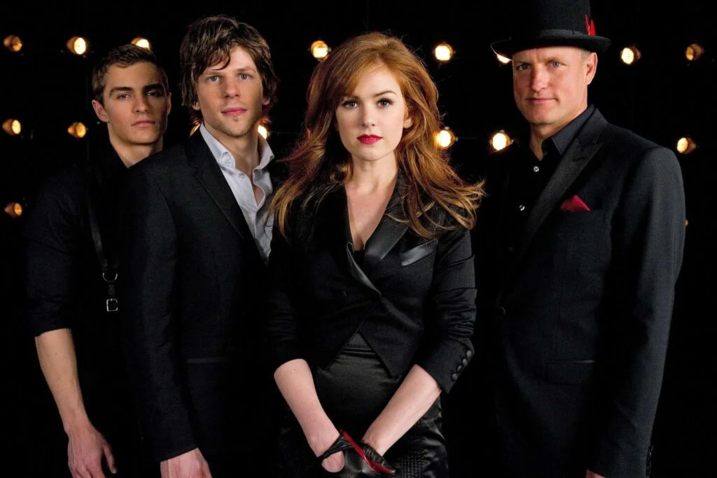 Four individuals dressed in black formal attire stand together against a dark backdrop with bright stage lights. The woman in front has long red hair and is wearing a tailored blazer, while the three men behind her have varying hairstyles and expressions, contributing to a mysterious and dramatic group pose.