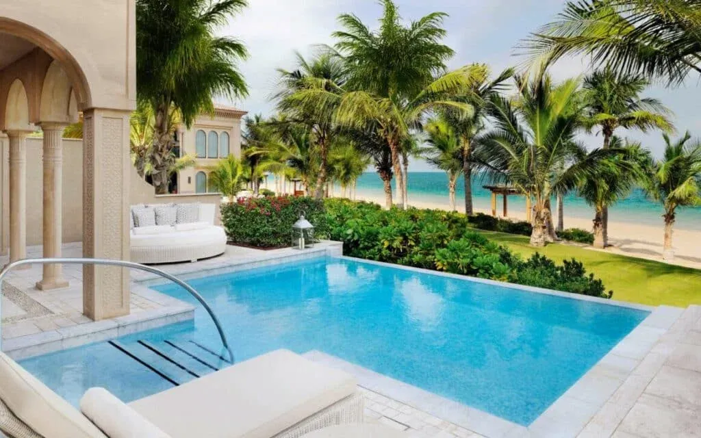 A luxurious poolside view surrounded by palm trees, with a well-maintained garden and beach visible in the background, showcasing a mix of greenery and the tranquil blue sea.