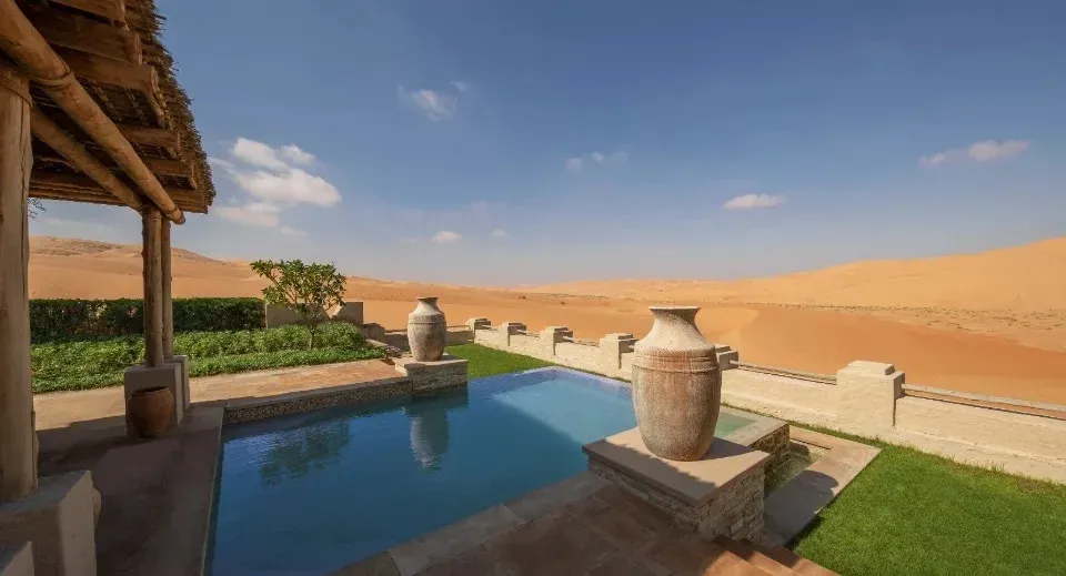 A luxurious villa patio featuring a swimming pool with two decorative stone vases, surrounded by plants and a view of expansive sand dunes under a blue sky with scattered clouds.