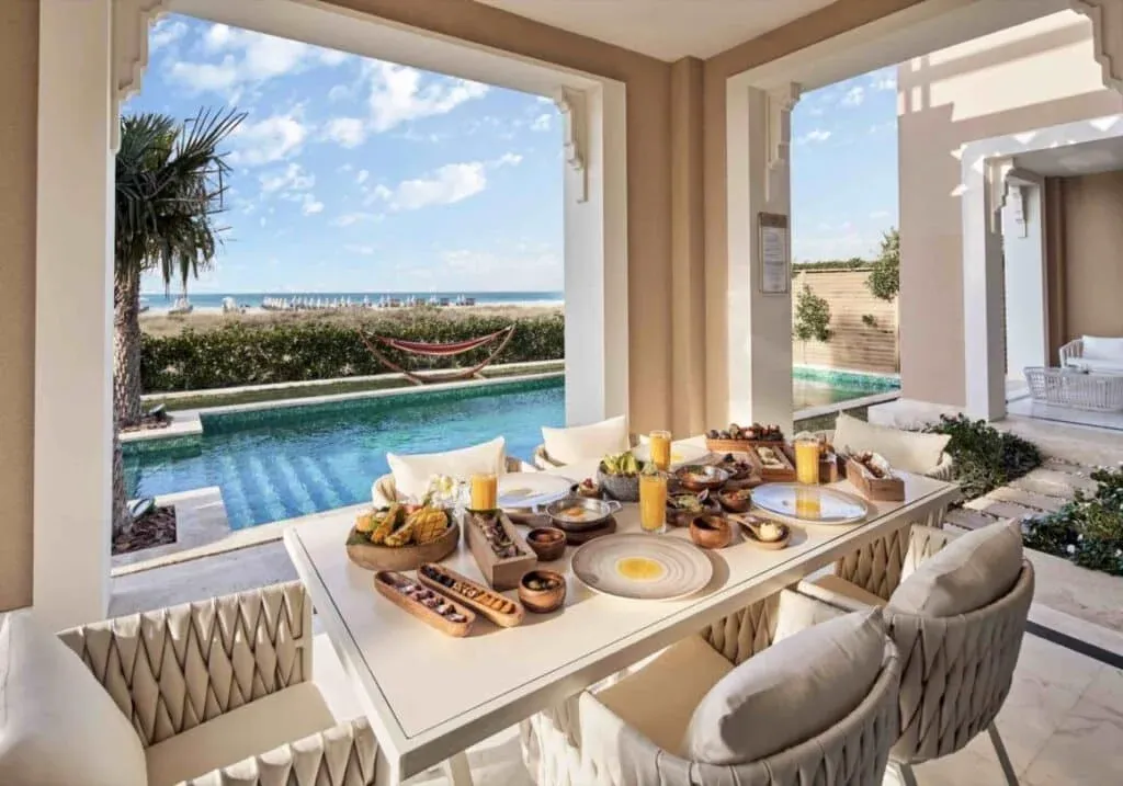 A beautifully set outdoor dining table featuring a variety of dishes and drinks, including tropical fruits and pastries, with a view of a swimming pool and the ocean. A hammock is visible in the background under clear blue skies.