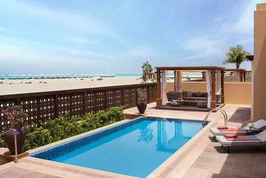 A luxurious outdoor space featuring a private swimming pool, comfortable seating area under a wooden pergola, and landscaped greenery, set against a backdrop of a sandy beach and clear blue ocean under a bright sky.