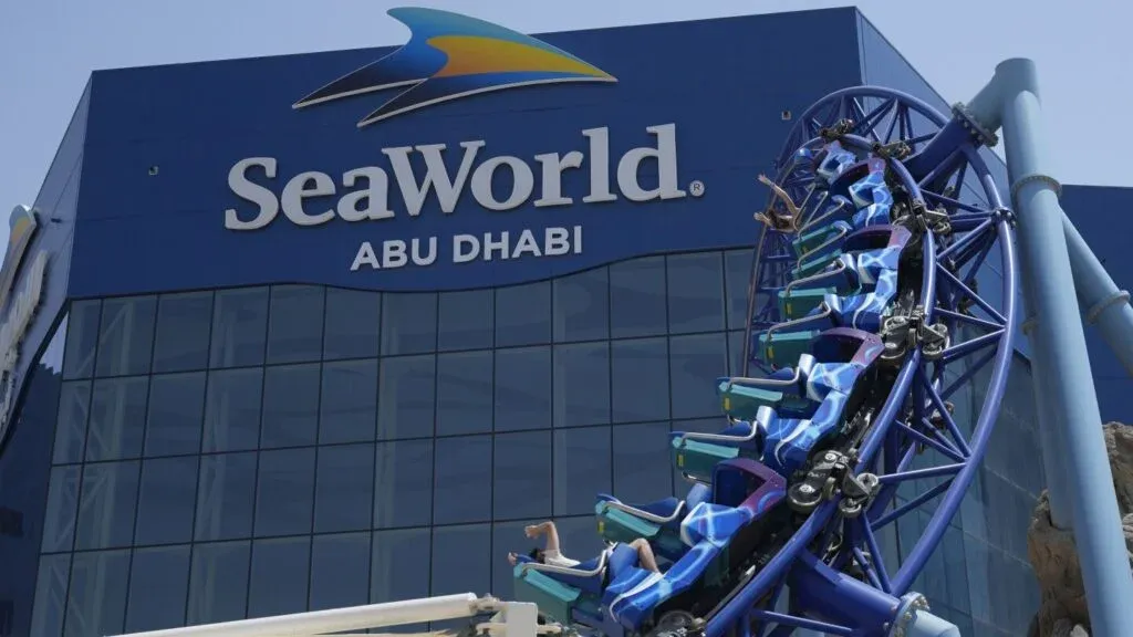 A roller coaster descends outside the SeaWorld Abu Dhabi building, which features a large blue sign with the SeaWorld logo and the name of the city. The roller coaster has blue and purple seats with riders visible, enjoying the thrill.
