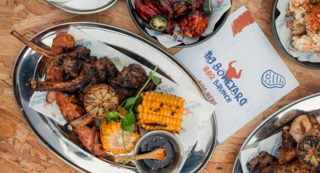 A silver platter featuring an assortment of grilled meats including ribs and chicken, alongside grilled corn and a small bowl of sauce, set on a wooden table with a restaurant menu partially visible.