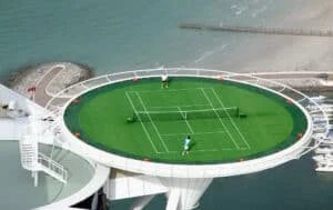 Aerial view of a round, rooftop tennis court on a building, surrounded by water and overlooking a sandy beach; two players are engaged in a match on the bright green court.