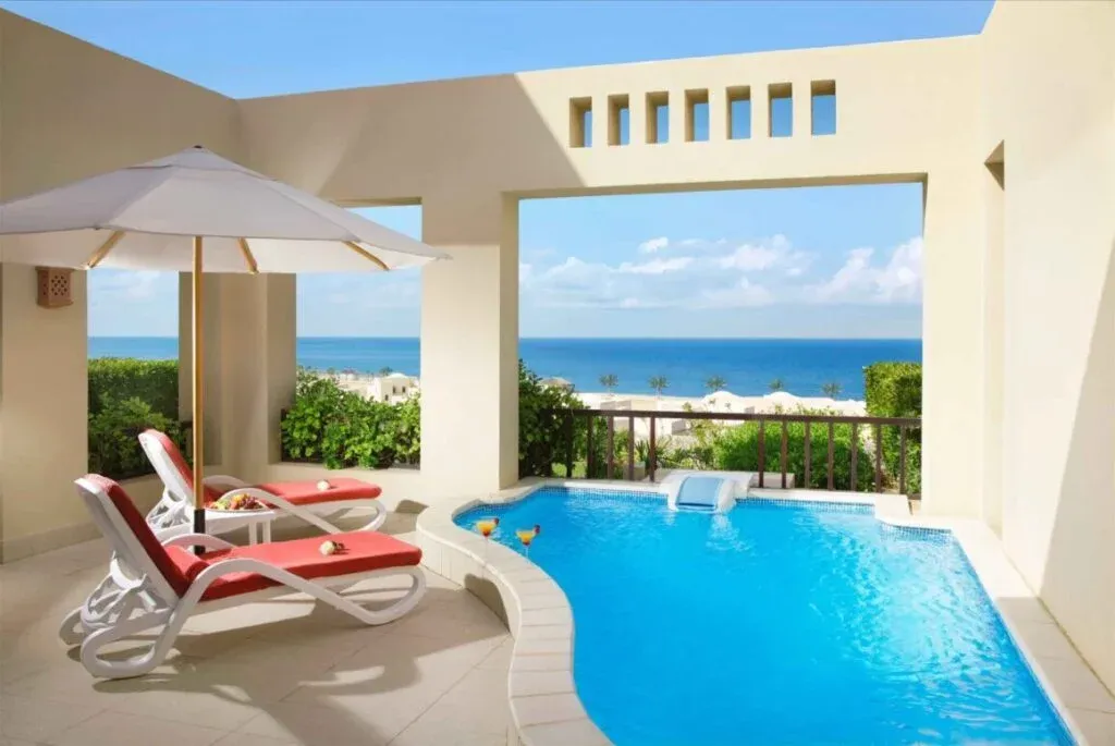 A luxurious outdoor space featuring two lounge chairs with red cushions, an umbrella, and a small table with fruit, adjacent to a private pool overlooking the ocean and palm trees under a blue sky.