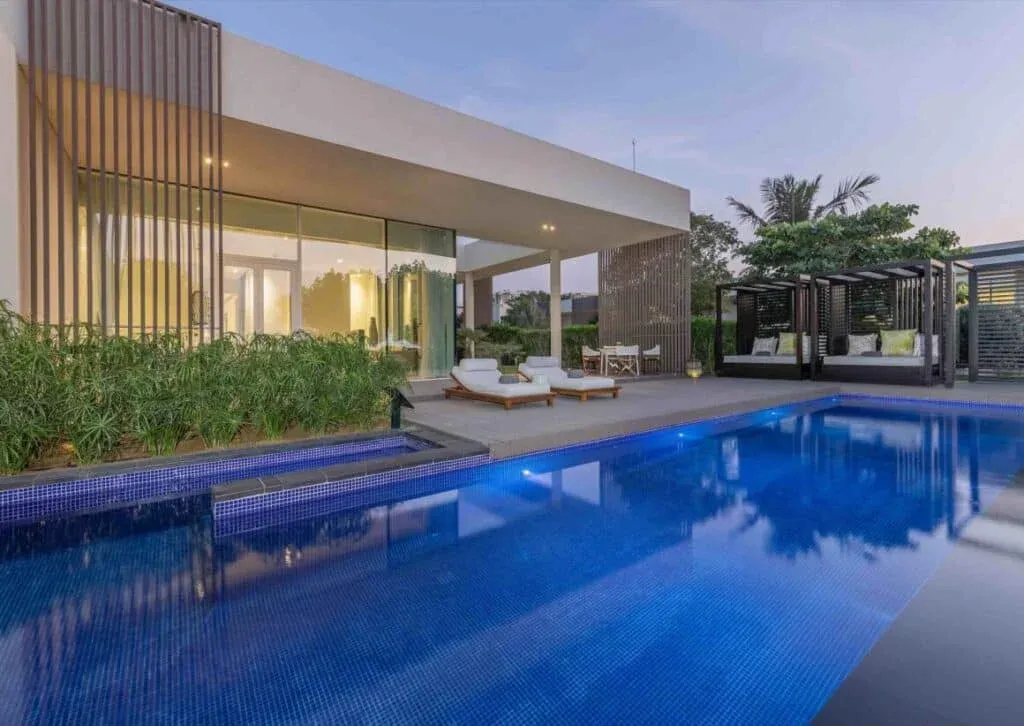 A modern house with large glass windows overlooking a blue swimming pool, featuring a deck with white lounge chairs and lush green plants nearby, and cool evening lighting under a dusky sky. A shaded seating area is visible in the background.