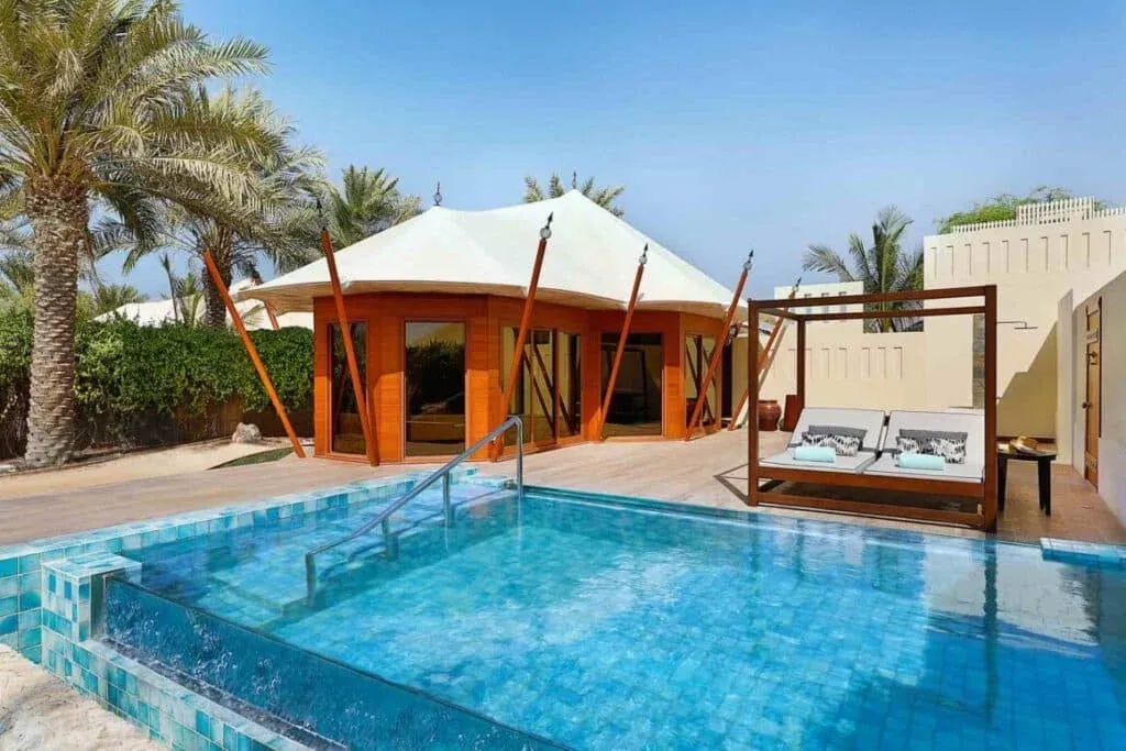 A luxurious pool area featuring a rectangular swimming pool with blue tiles, surrounded by palm trees. In the background, there is a wooden structure with large windows and a tan canvas roof. A stylish daybed with soft cushions is positioned beside the pool, all set against a clear blue sky.