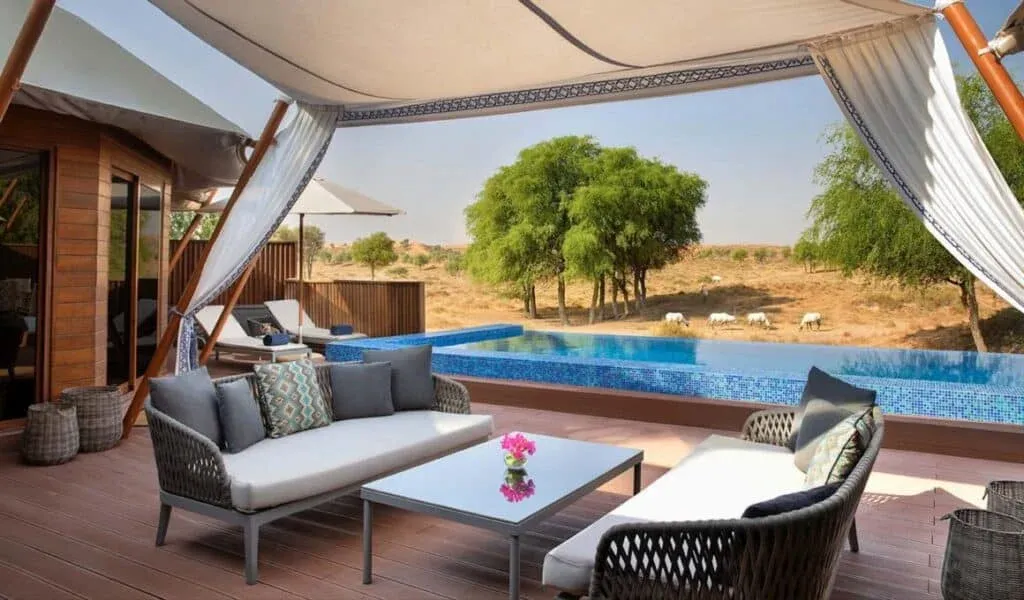 A stylish outdoor lounge area features a white sofa adorned with colorful cushions and a glass coffee table with a small flowering plant, overlooking a serene swimming pool and a landscape with green trees and distant grazing cattle under a clear sky.