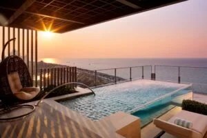 A serene balcony view featuring a modern infinity pool overlooking the ocean at sunset, with a hanging wicker chair and a minimalist lounge chair with a rolled towel, all set against a backdrop of vibrant orange and purple sky.